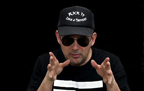 DJ Westbam wearing a cap and shades in front of black background.
