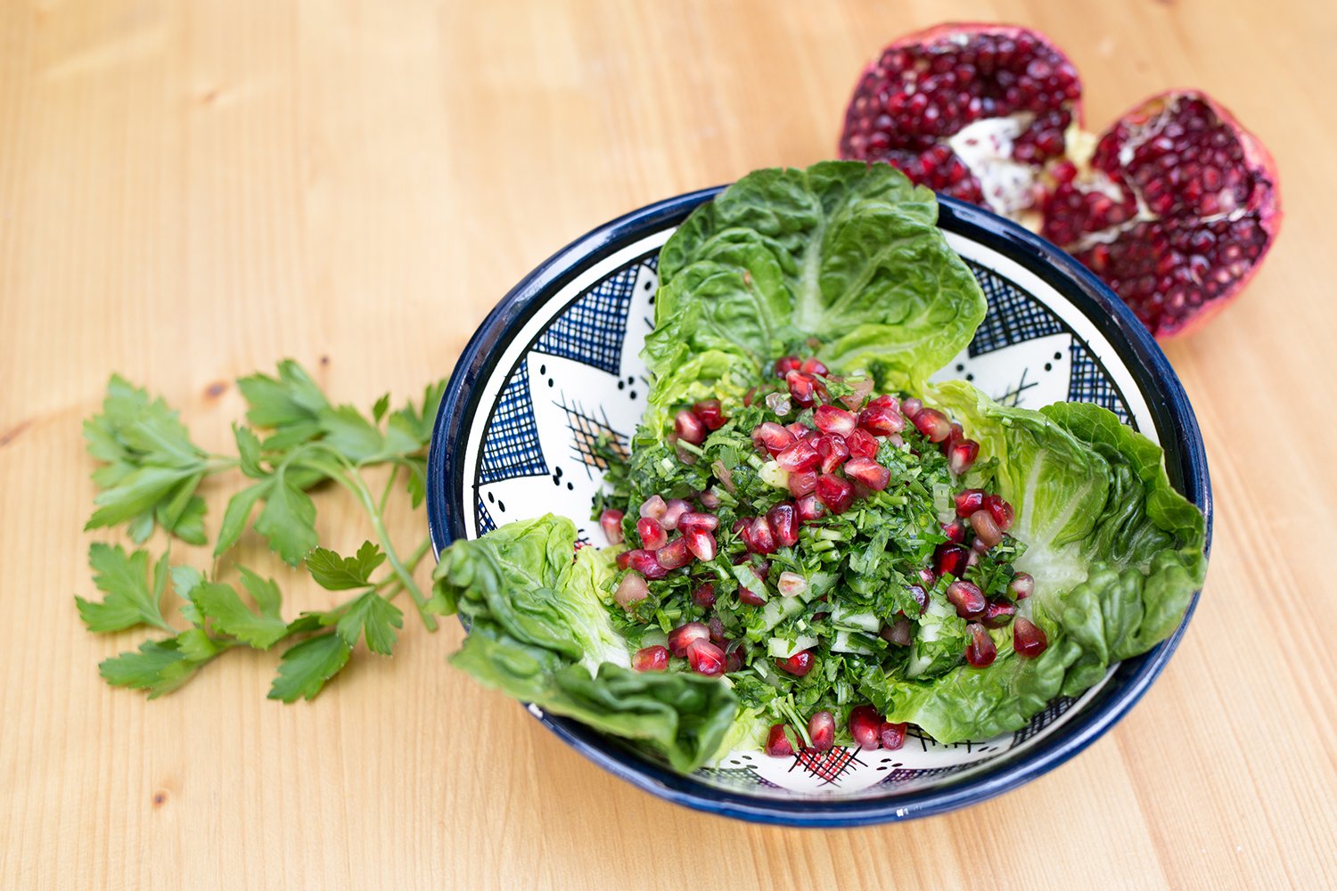 Salad in blue-white patterned bowl with pomegranate seed on wooden table. Pomegranate and Koriander in background. A dish at Friedrichshain restaurant Aleppo Supper Club.