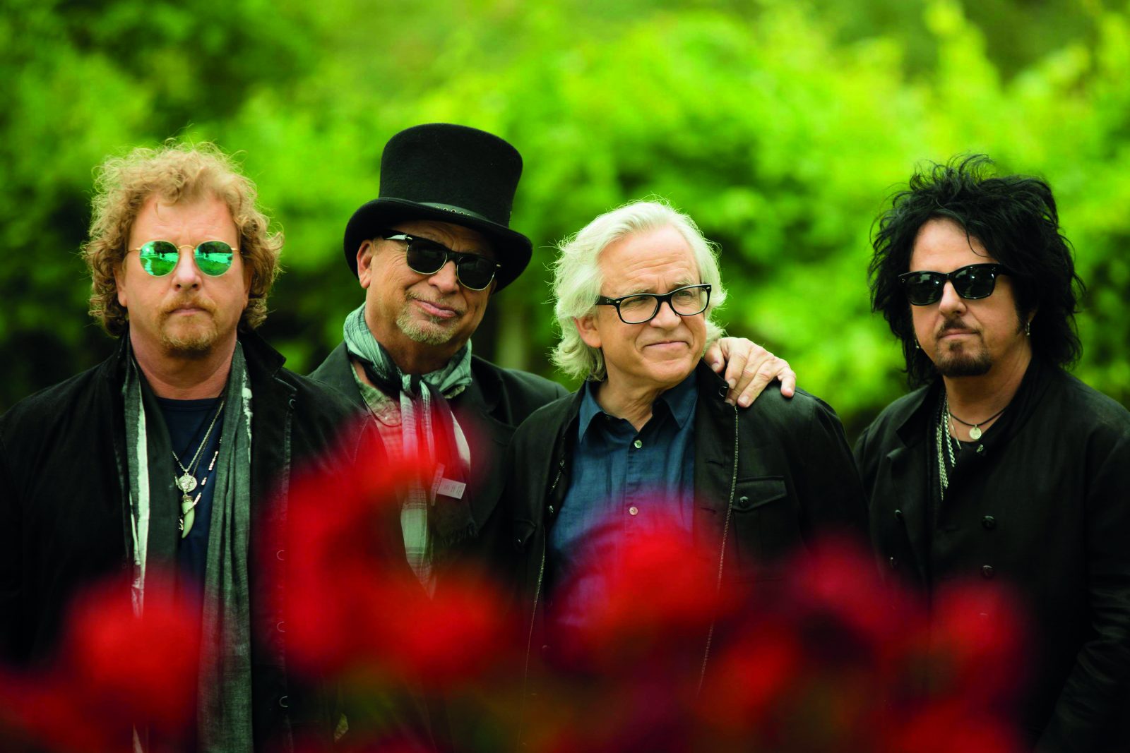 Die Band Toto, vier ältere Männer in schwarzer Kleidung in einem Park, im Vordergrund rote Blumen.