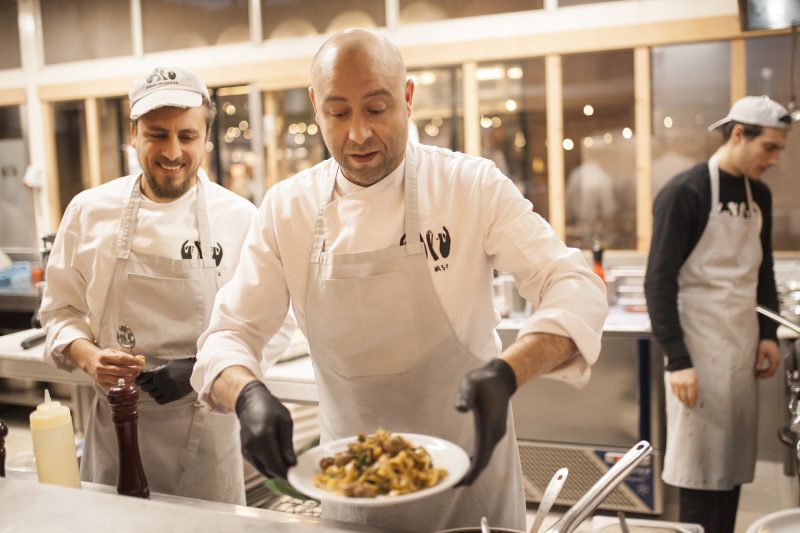 Ein Mann in Küche mit schwarzen Handschuhen serviert Teller Pasta auf Theke, zwei Männer im Hintergrund. Das Restaurant Mani in Pasta in Kreuzberg ist zwar geschlossen, die Manufaktur in der Markthalle Neun aber noch geöffnet.