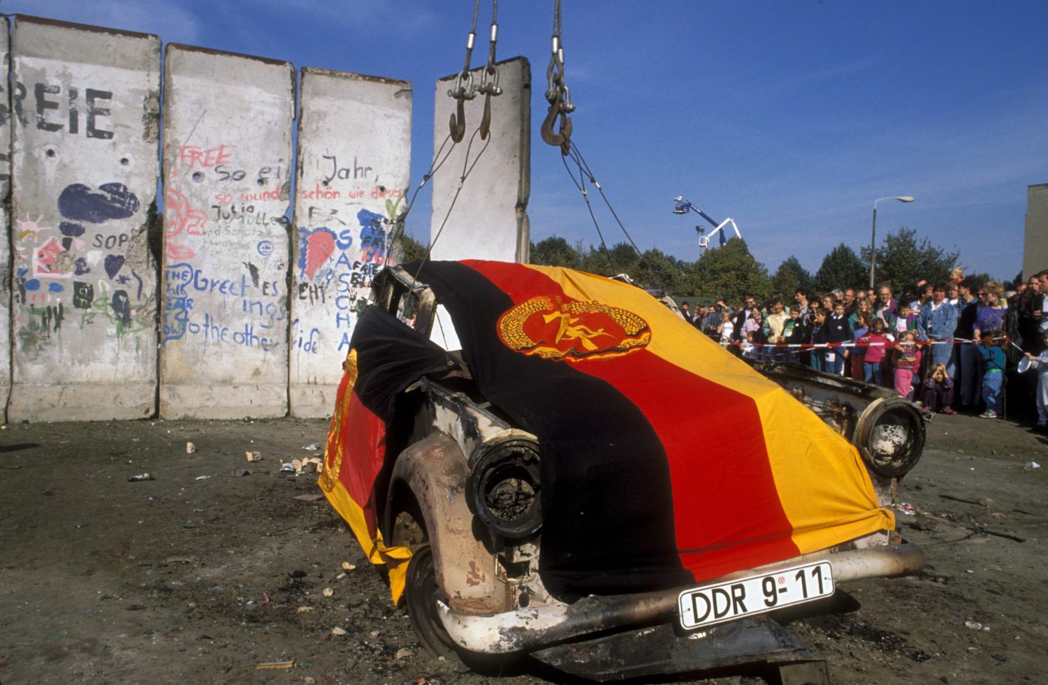 3. Oktober 1990: Verkohltes Wrack eines Trabanten geschmückt mit der DDR Fahne wird vor Resten der Berliner Mauer aufgestellt. Foto: Imago/Sven Simon
