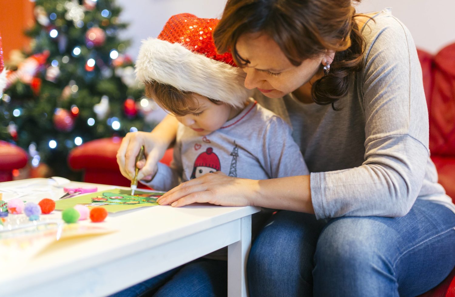 Eine Frau sitzt mit einem Kleinkind an einem Tisch und bastelt eine Weihnachtskarte. 
